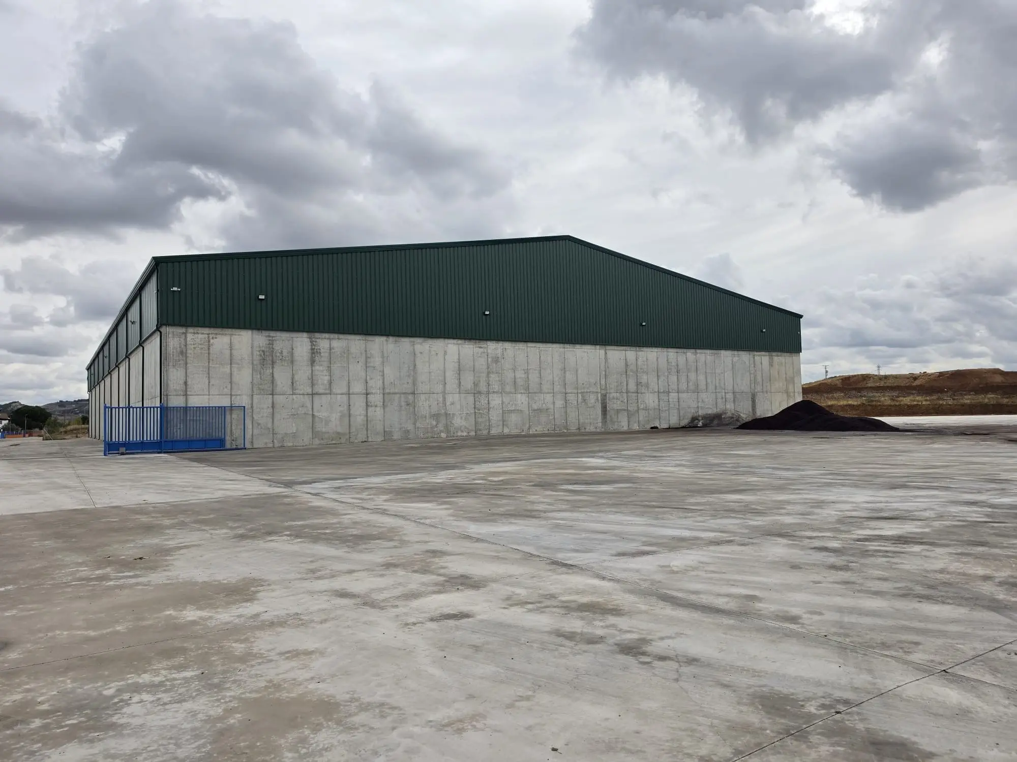 Nave de muros de hormigón y cubierta verde rodeada por una explanada de hormigón nueva