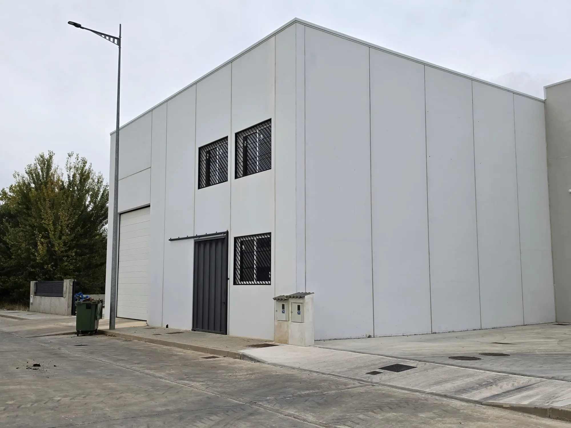 Nave de paneles blancos con ventanas enrejadas y portón, junto a una acera nueva