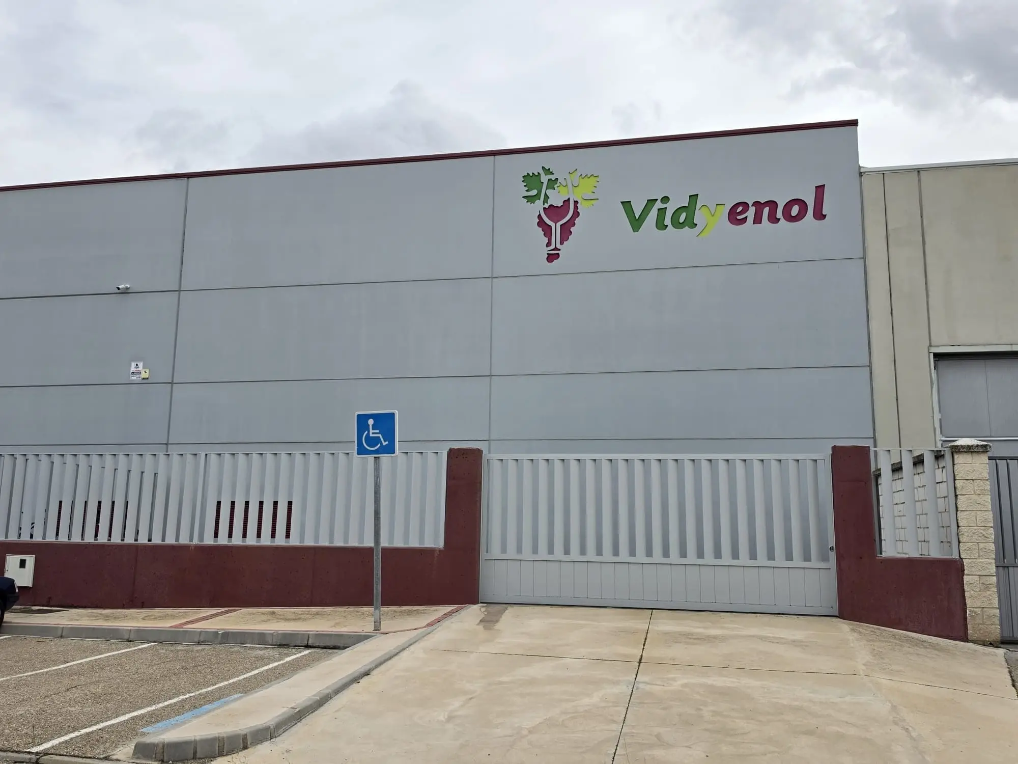 Fachada de paneles grises de la nave de Vidyenol con su logotipo y el aparcamiento delante