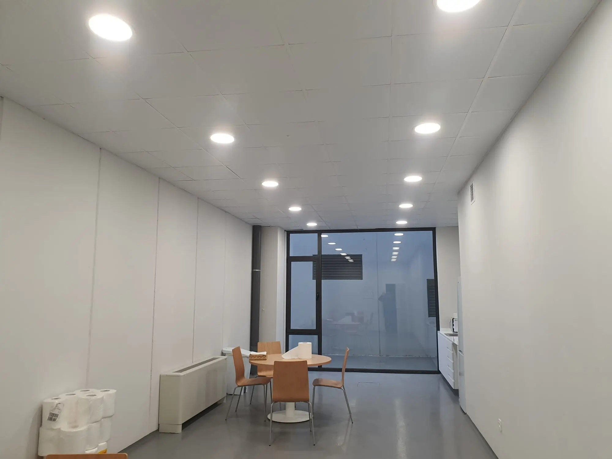Sala de oficinas con mesa redonda, techo de focos y puerta de cristal al fondo
