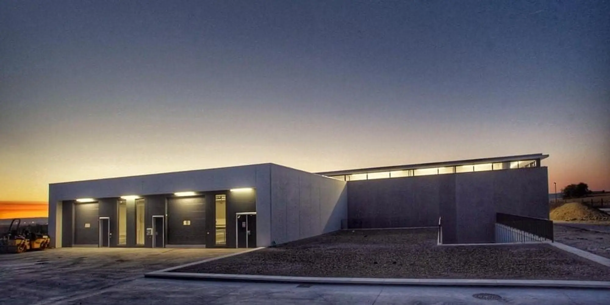 Bodega terminada al anochecer: edificio bajo y alargado con la franja de ventanas iluminada