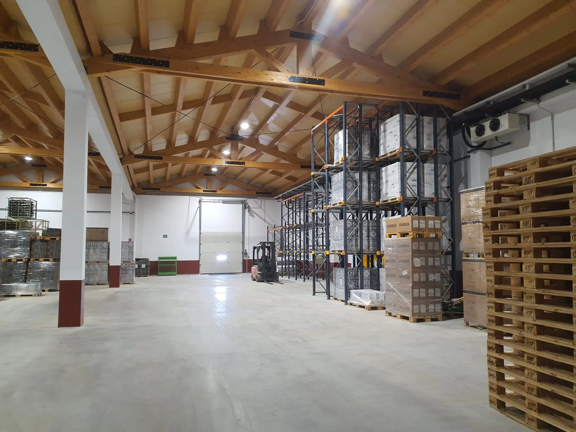 Nave con cerchas de madera laminada y pilares blancos, con estanterías y palés de botellas