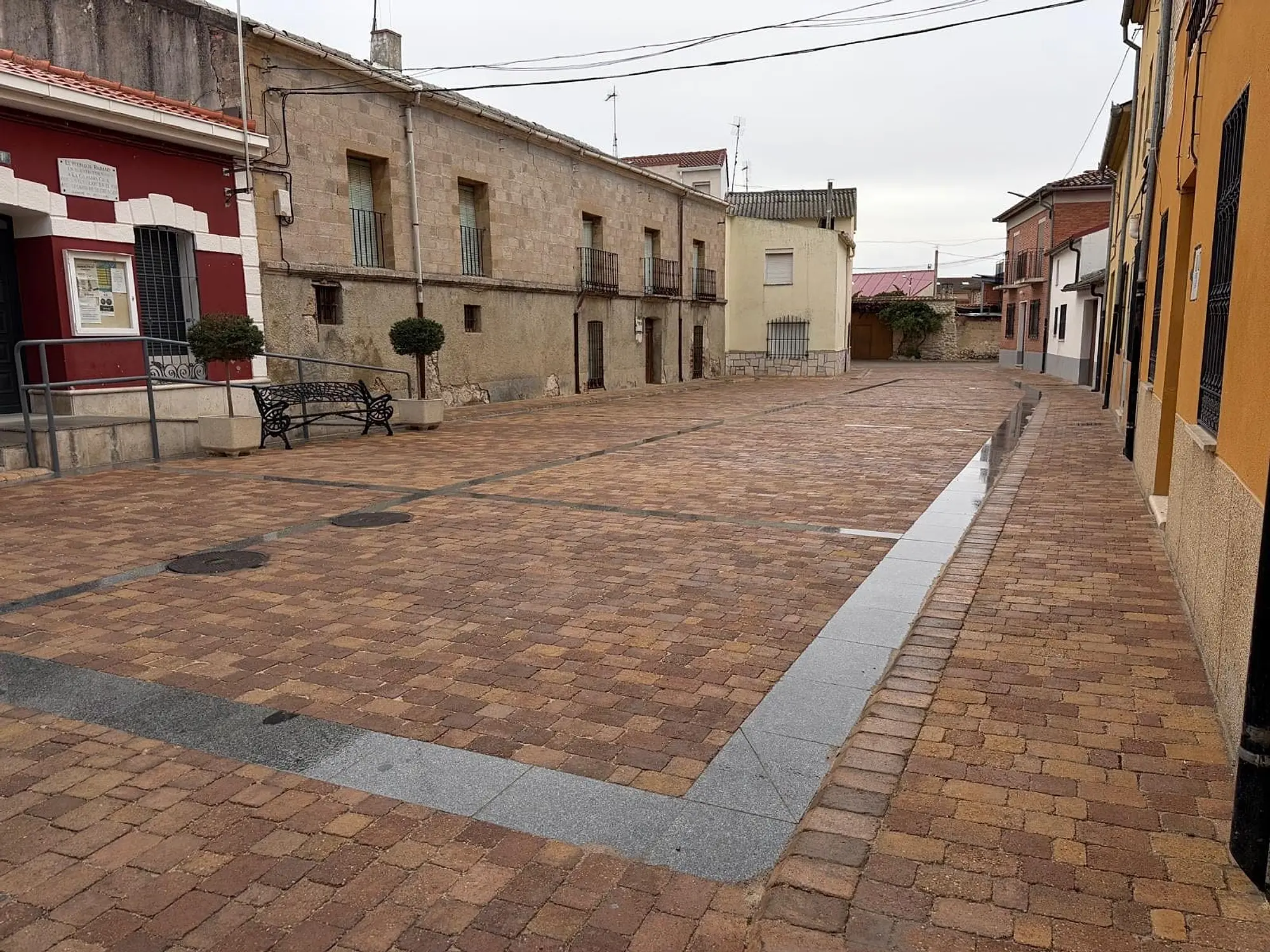 Plaza de Rábano con adoquín en tonos tierra, un banco y jardineras, y una banda de piedra gris junto a las casas