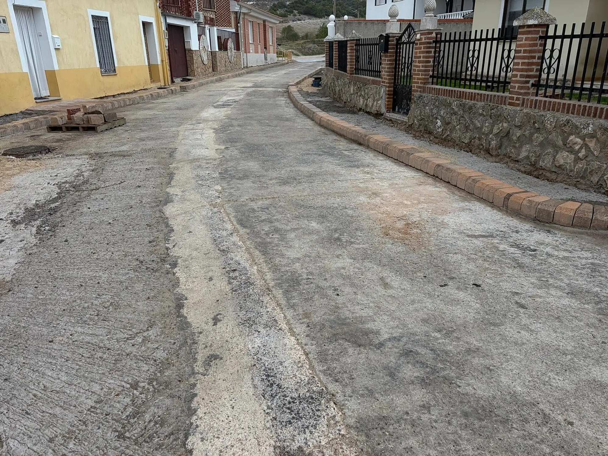 Calle en obra con los bordillos nuevos de la acera junto a una verja y un muro de piedra