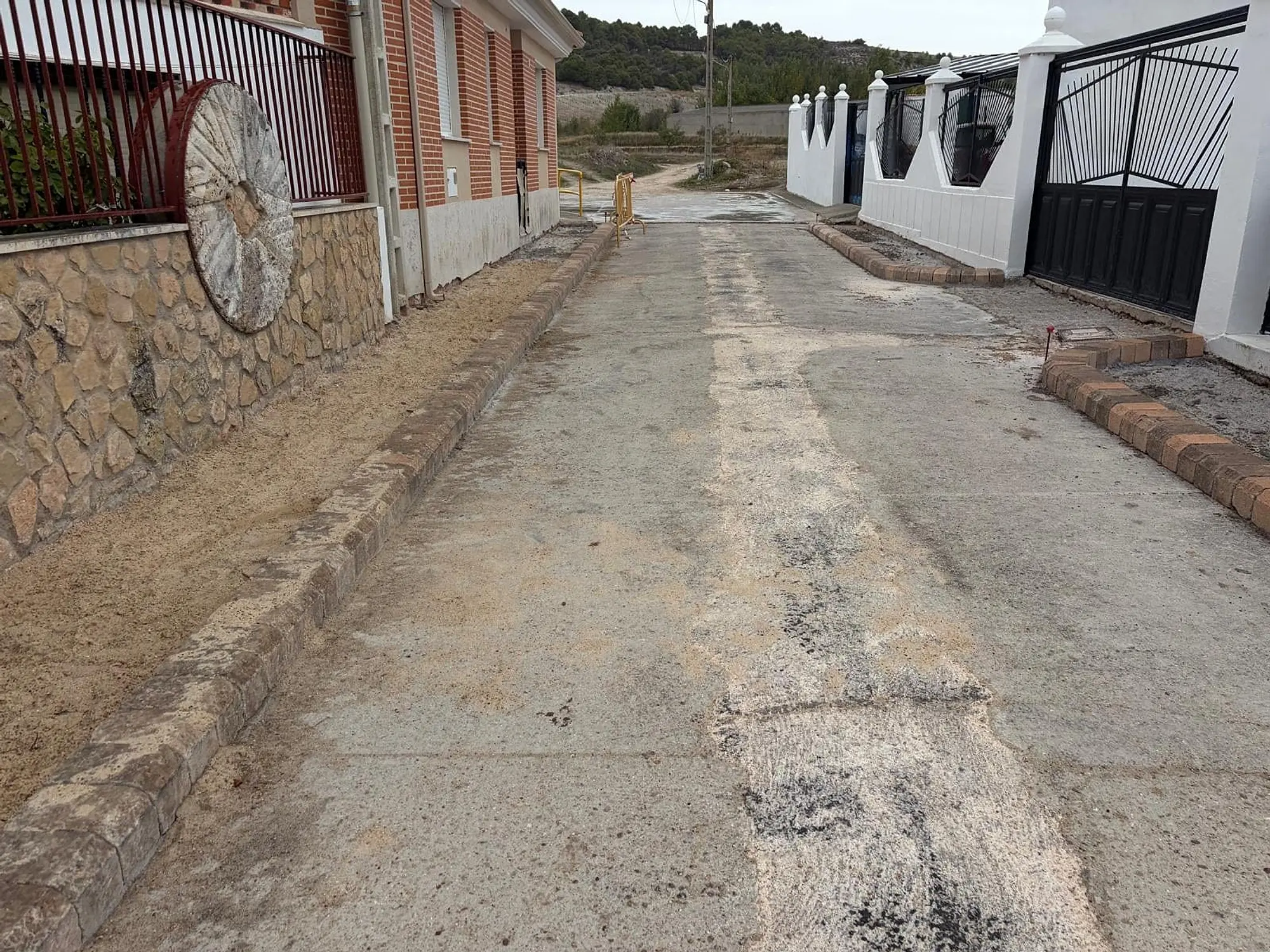 Calle en obra con bordillos nuevos entre una fachada con una rueda de carro y una verja blanca